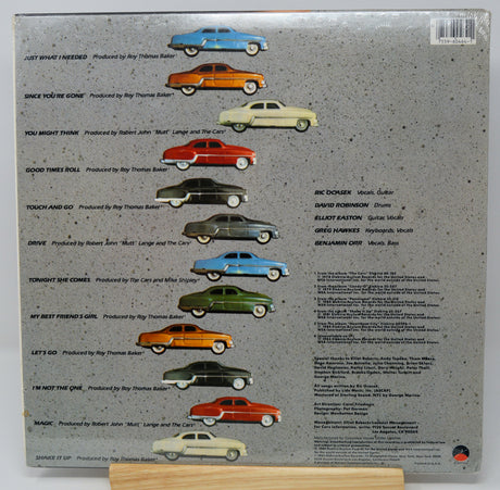 The back cover of a vinyl record sleeve for Ray Thomas Ballard's album 'Just What I Need', displayed on a small wooden stand. The cover features a textured gray background with a row of illustrated vintage cars in various colors (blue, orange, yellow, red, green, black, white) above a tracklist and credits.