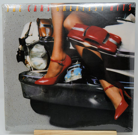 The Cars Greatest Hits vinyl record cover, sealed in plastic shrink wrap and displayed on a small wooden stand. The cover art features a woman's legs in red Mary Jane heels sitting on a motorcycle, with a red car graphic overlay and the band's name in orange text at the top.