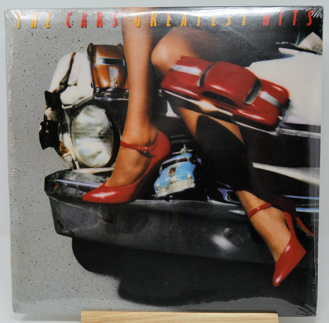 The Cars Greatest Hits vinyl record cover, sealed in plastic shrink wrap and displayed on a small wooden stand. The cover art features a woman's legs in red Mary Jane heels sitting on a motorcycle, with a red car graphic overlay and the band's name in orange text at the top.