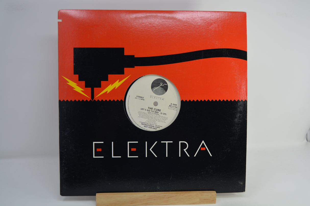 The album cover of The Cure's 'Let's Go To Bed' is displayed on a small wooden stand against a white background. The cover features a vibrant, multicolored geometric border