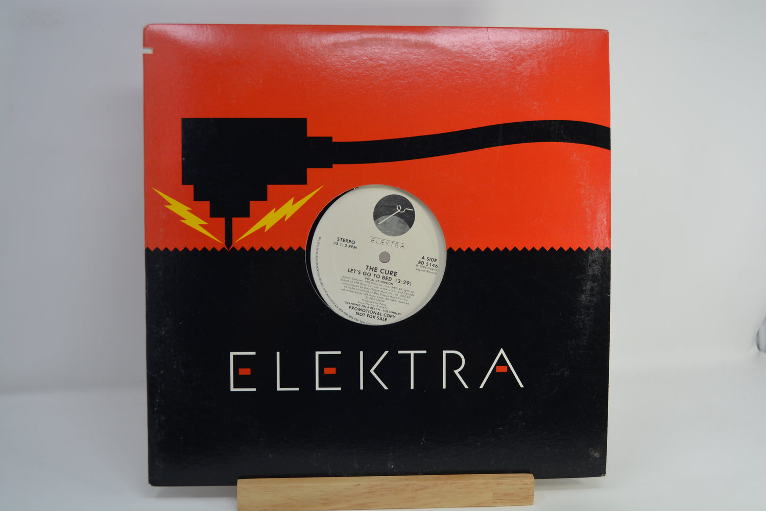 The album cover of The Cure's 'Let's Go To Bed' is displayed on a small wooden stand against a white background. The cover features a vibrant, multicolored geometric border