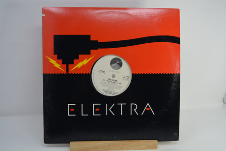 The album cover of The Cure's 'Let's Go To Bed' is displayed on a small wooden stand against a white background. The cover features a vibrant, multicolored geometric border