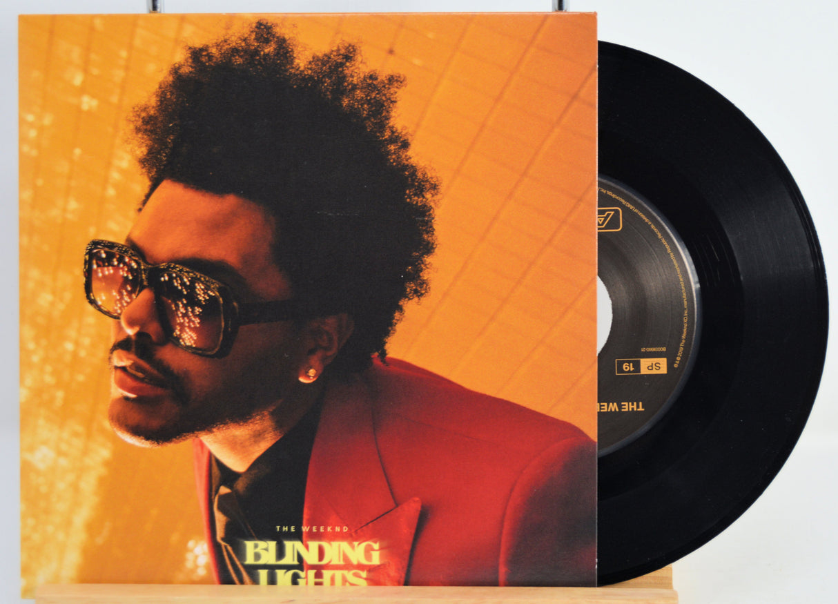 Vinyl record with a cover featuring The Weeknd wearing sunglasses and a red jacket, set against an orange and gray gradient background. Song - Blinding Lights