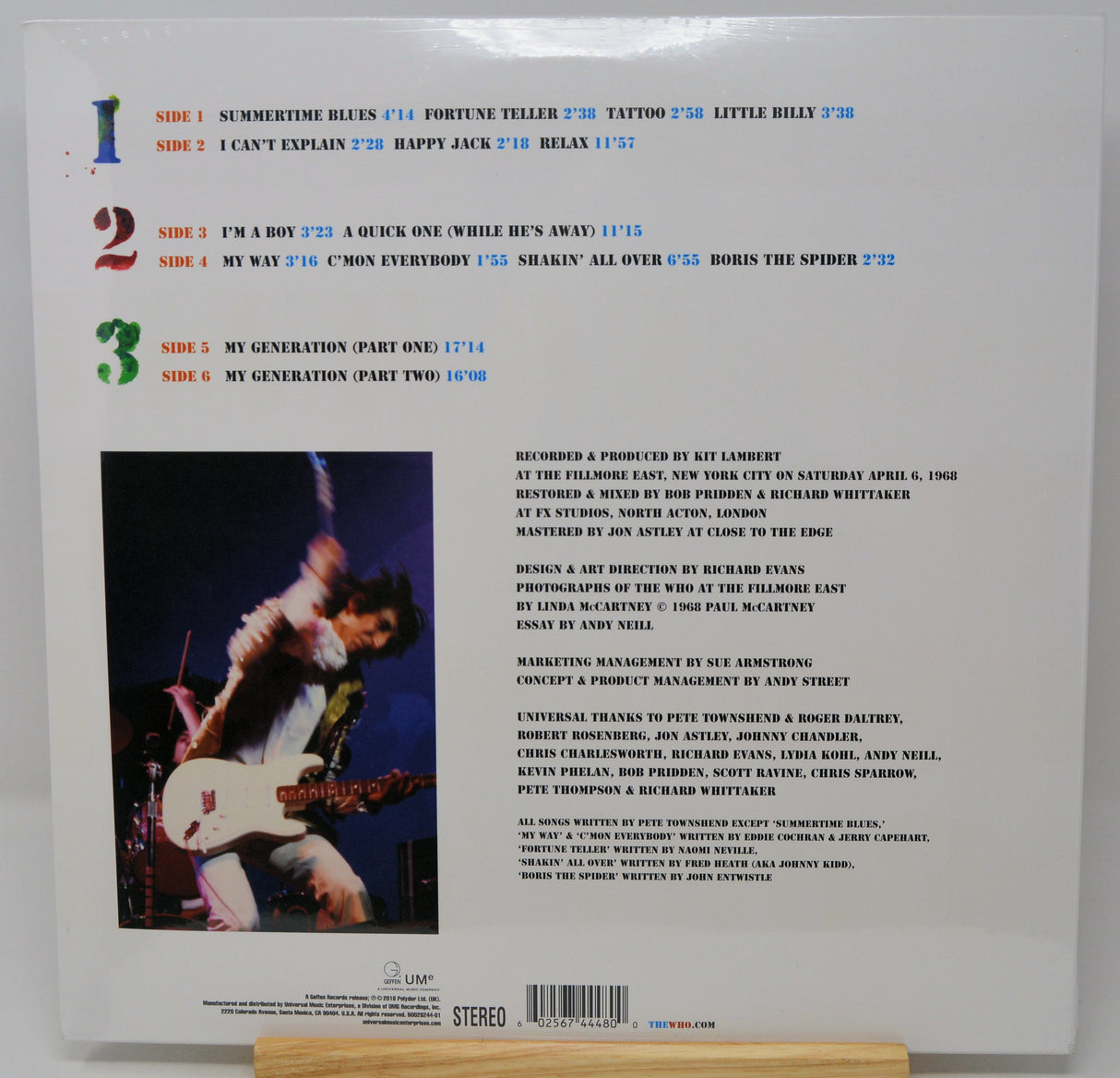 The back cover of a vinyl record sleeve displayed on a small wooden stand. The cover is white with multicolored track numbers (blue, red, green, purple), black text, a small black-and-white photo of a hand playing a guitar, and a barcode at the bottom.