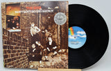 Vinyl record with cover featuring a group of people on a brick wall.
