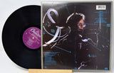 A vinyl record partially pulled from its album sleeve, showing the black record and a purple center label next to the printed back cover artwork with blue line art and a portrait.