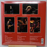 The back cover of a vinyl record sleeve, featuring a solid red background with five black-and-white concert photos of musicians, track listings, and credits on a wooden display stand.