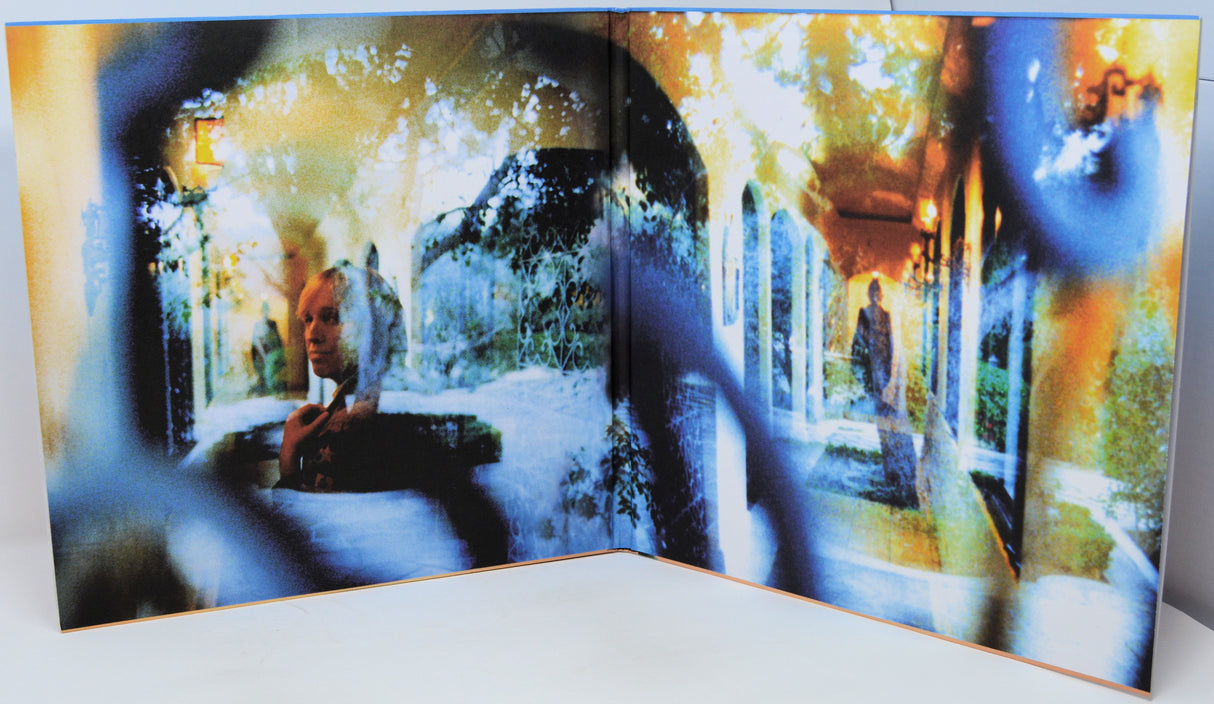 An open art book or photo print set displayed on a white surface, showing a surreal, double-exposure style image with a seated woman on the left page and a standing figure in a corridor on the right page, overlaid with blue and yellow abstract visual effects.