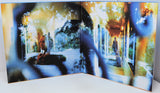 An open art book or photo print set displayed on a white surface, showing a surreal, double-exposure style image with a seated woman on the left page and a standing figure in a corridor on the right page, overlaid with blue and yellow abstract visual effects.