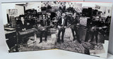 A black and white photographic print or poster showing a band of five musicians in a gear-filled studio, displayed on a white surface.