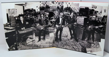 A black and white photographic print or poster showing a band of five musicians in a gear-filled studio, displayed on a white surface.