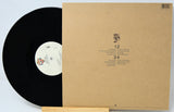 A vinyl record partially pulled from its kraft paper sleeve, displayed on a small wooden stand against a white background.