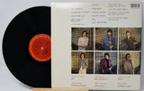 A vinyl record partially pulled from its back cover sleeve, showing the black record on the left and the printed back of the album case on the right. The sleeve includes a barcode, tracklist, handwritten notes, and six small group portraits.