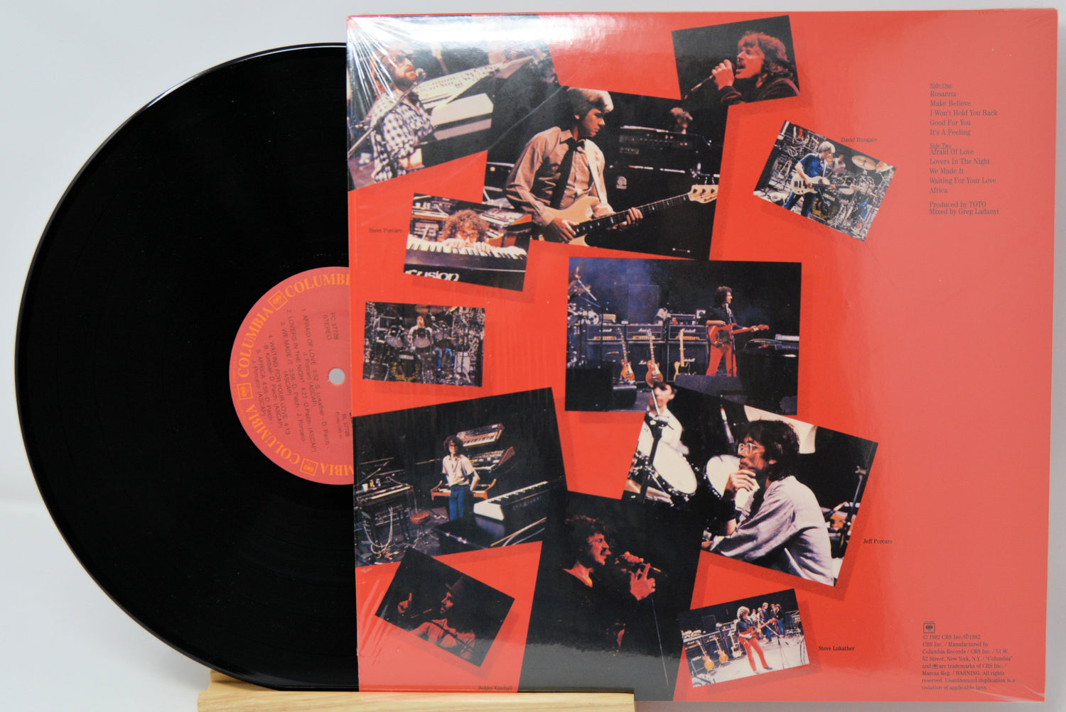 A vinyl record partially pulled from its sleeve, showing a black record on the left and the album cover on the right. The sleeve has a bright red-orange background with a collage of small black-and-white and color photos of a musician performing, plus tracklist text on the right side.