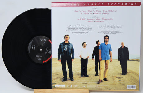 A vinyl record partially pulled from its back cover sleeve, showing the black record on the left and the printed back cover on the right. The cover features a group photo of four men standing outdoors on a sandy landscape, with track listing text at the top and a red header reading 'ORIGINAL MASTER RECORDING'.