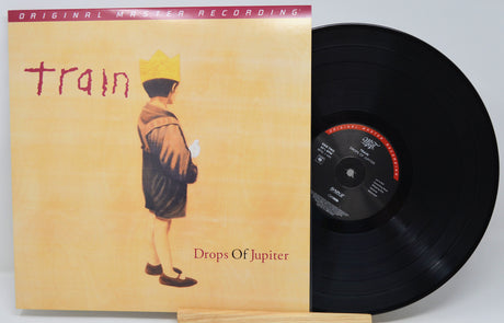 Train's 'Drops Of Jupiter' original master recording vinyl LP, showing the illustrated album cover sleeve next to the black 12-inch record on a stand.