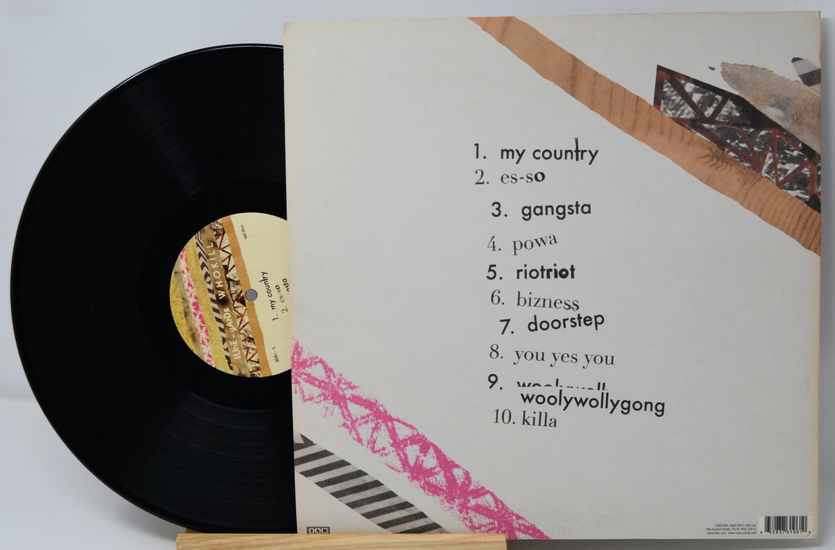 A vinyl record partially pulled from its sleeve, showing the tracklist cover insert. The black record has a patterned label, and the sleeve is off-white with black text listing ten tracks and abstract pink, brown, and black graphic accents.