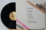 A vinyl record partially pulled from its sleeve, showing the tracklist cover insert. The black record has a patterned label, and the sleeve is off-white with black text listing ten tracks and abstract pink, brown, and black graphic accents.