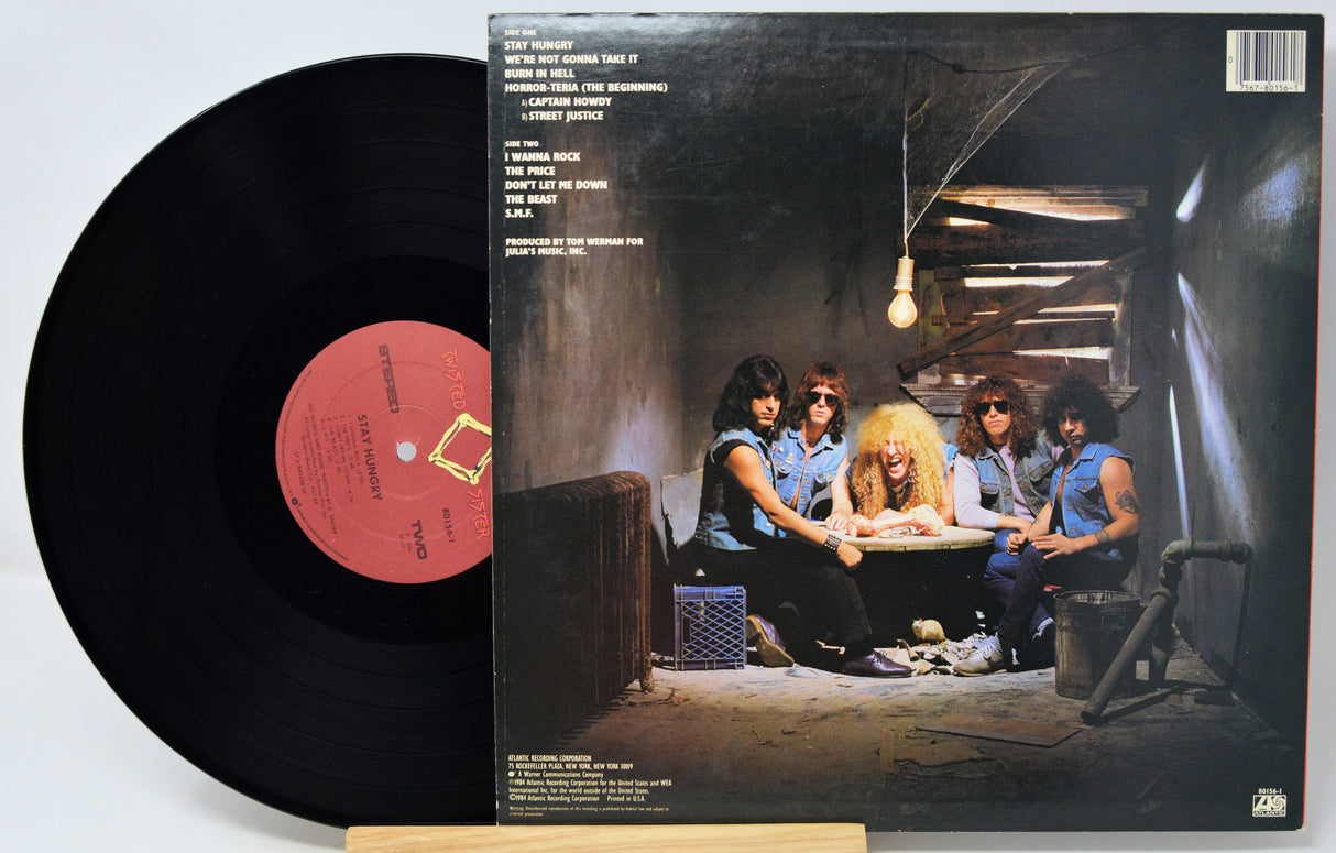 A vinyl record partially pulled from its album sleeve, showing the back cover artwork of a band in a dim interior scene. The black record has a pink-red center label with printed text.