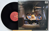A vinyl record partially pulled from its album sleeve, showing the back cover artwork of a band in a dim interior scene. The black record has a pink-red center label with printed text.