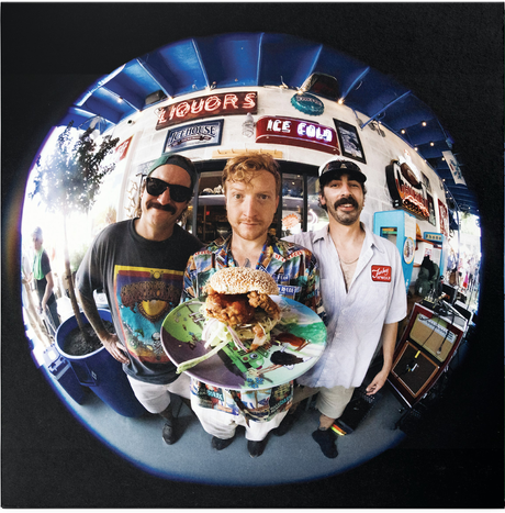 Three men in a fast-food restaurant setting with a large burger graphic on a plate.