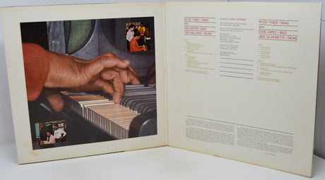An open vintage music book or album booklet showing printed sheet music or lyrics on the right page and a large inset photograph of hands playing a piano on the left page.