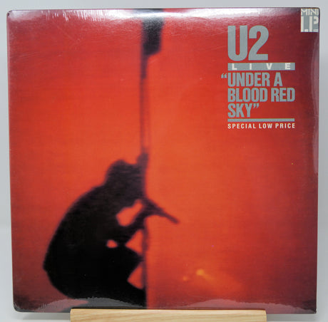 U2 'Under a Blood Red Sky' live vinyl record LP cover, sealed in plastic shrink wrap and displayed on a small wooden stand against a light gray background.