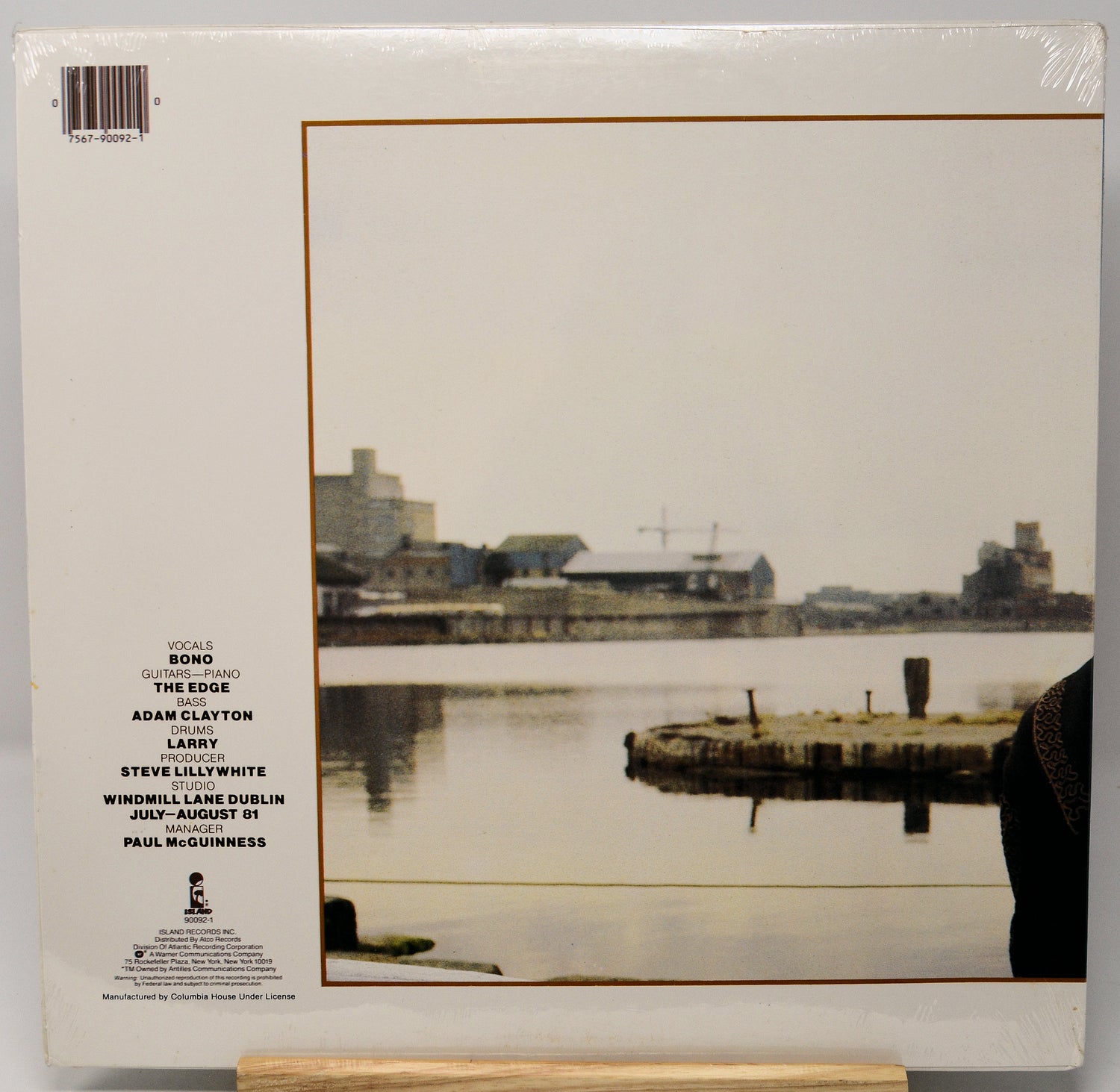 The back cover of a vinyl record sleeve, sealed in plastic shrink wrap, featuring a photograph of a waterfront scene with industrial buildings and a pier. The left side lists song titles and artist names including Adam Clayton, Larry, Steve Lillywhite, and Paul McGuinness.