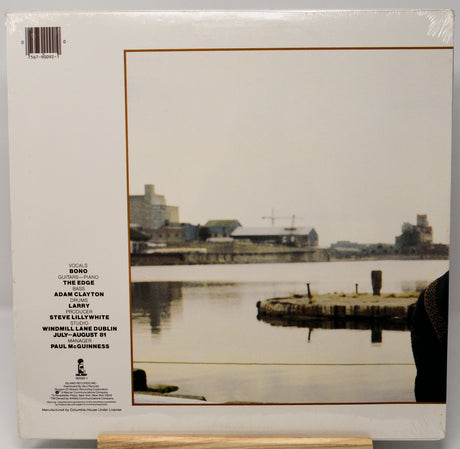The back cover of a vinyl record sleeve, sealed in plastic shrink wrap, featuring a photograph of a waterfront scene with industrial buildings and a pier. The left side lists song titles and artist names including Adam Clayton, Larry, Steve Lillywhite, and Paul McGuinness.