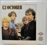 A U2 'October' vinyl record LP in its original shrink wrap, displayed on a small wooden stand against a white background.