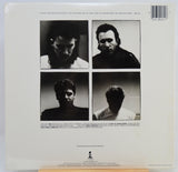 The back cover of a vinyl record sleeve  featuring four black and white portrait photographs of the band members. The cover is printed on white paper and is sealed in plastic shrink-wrap, resting on a light-colored wooden surface.