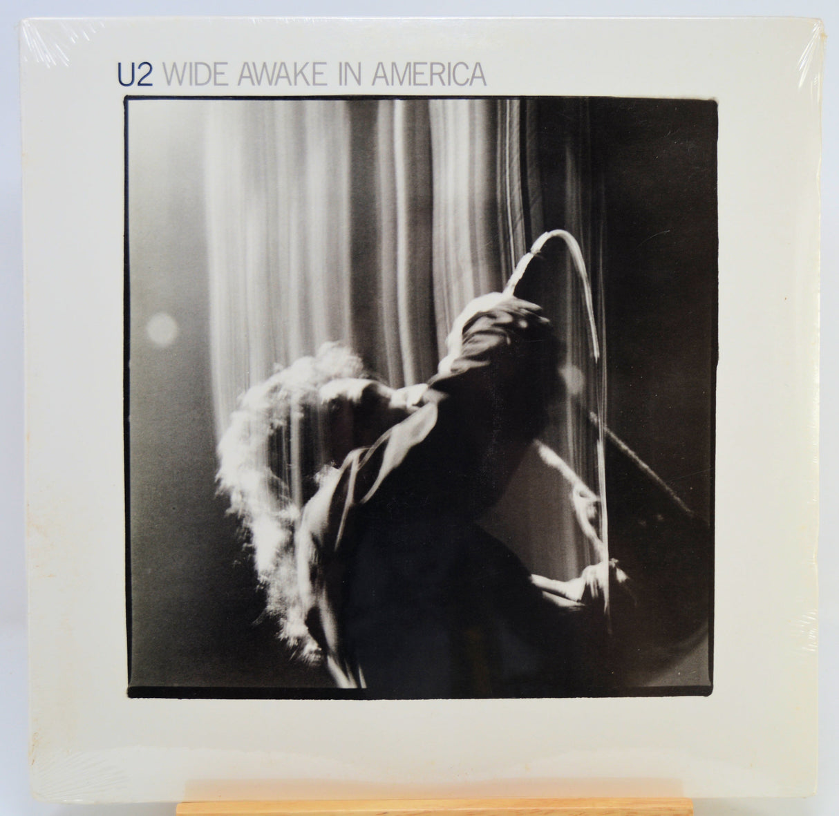 U2 'Wide Awake in America' vinyl record album cover, sealed in plastic, displayed on a wooden stand against a white background.
