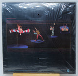The back cover of a vintage vinyl record sleeve, sealed in its original plastic shrink wrap. The cover is predominantly black with a central graphic depicting three performers on stage: a trapeze artist, a guitarist, and a singer on a microphone stand, surrounded by extensive white and light blue text credits.