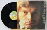 A vinyl record partially pulled from its album cover, showing a close-up portrait of a man on the cover and a black record disc with a colorful center label.