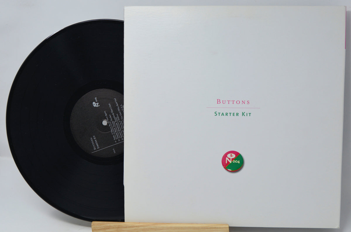 A vinyl record partially pulled from its white sleeve, displayed on a small wooden stand against a light gray background. The sleeve reads 'Buttons Starter Kit' with a small circular red and green logo near the bottom.