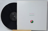 A vinyl record partially pulled from its white sleeve, displayed on a small wooden stand against a light gray background. The sleeve reads 'Buttons Starter Kit' with a small circular red and green logo near the bottom.