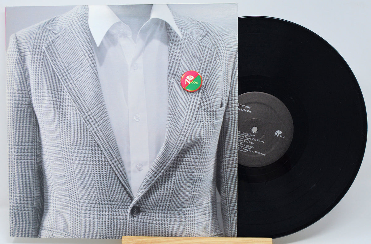 A vinyl record partially covering a printed sleeve or cover art showing a mannequin torso in a light gray plaid blazer over a white shirt with a small red and green pin at the chest. The black record is visible on the right against a light gray background.