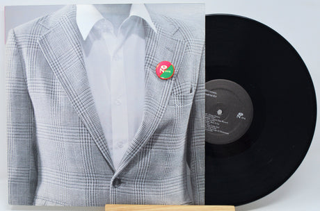 A vinyl record partially covering a printed sleeve or cover art showing a mannequin torso in a light gray plaid blazer over a white shirt with a small red and green pin at the chest. The black record is visible on the right against a light gray background.