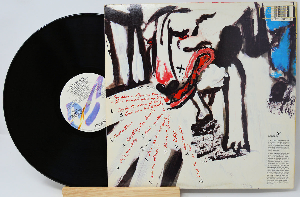 A vinyl record partially pulled from its illustrated album cover, displayed on a small wooden stand against a light gray background. The cover features an abstract black, white, and red painting with handwritten red text, and the black record has a white center label.
