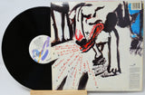 A vinyl record partially pulled from its illustrated album cover, displayed on a small wooden stand against a light gray background. The cover features an abstract black, white, and red painting with handwritten red text, and the black record has a white center label.