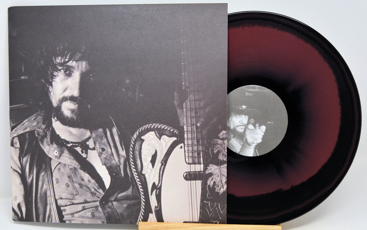 A vinyl record partially pulled from its sleeve, showing a black-and-white album cover with a bearded man holding a guitar and a maroon-to-black gradient record on a white background.