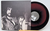 A vinyl record partially pulled from its sleeve, showing a black-and-white album cover with a bearded man holding a guitar and a maroon-to-black gradient record on a white background.