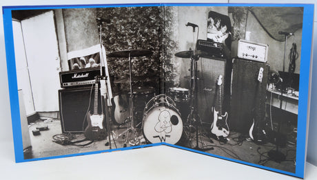 A square art print or poster with a vivid blue border, featuring a black and white photograph of a music studio setup with guitars, amplifiers, a drum kit, and a microphone stand.