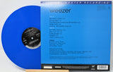 Weezer 'Blue Album' vinyl record shown with the blue LP partially pulled from the black-and-white inner label, alongside the blue printed album cover. Visible text includes 'weezer' and 'Original Master Recording.