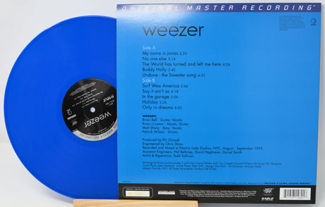 Weezer 'Blue Album' vinyl record shown with the blue LP partially pulled from the black-and-white inner label, alongside the blue printed album cover. Visible text includes 'weezer' and 'Original Master Recording.