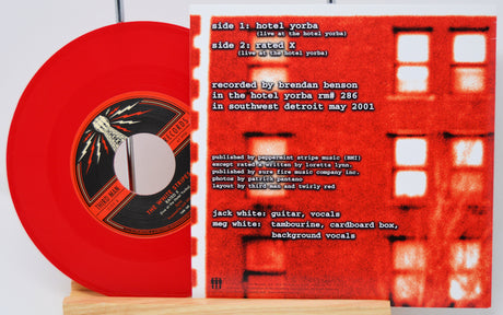 A red vinyl record partially pulled from its sleeve, displayed on a wooden stand. The sleeve is bright red with white text listing track titles and credits, including 'hotel yorba' and 'race x', with a faint architectural window pattern in the background.