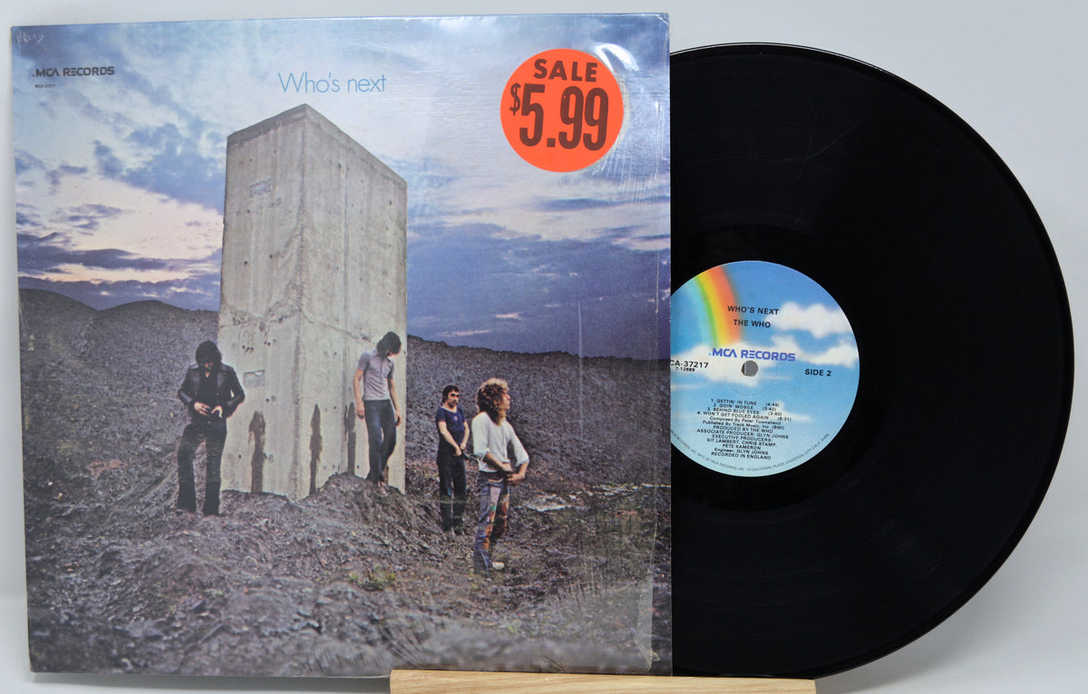 A vinyl record and its album cover for 'Who's next' by Axl Records, displayed on a small wooden stand against a light gray background. The cover features a desert landscape with a large concrete structure and three figures under a cloudy sky, with a red 'SALE $5.