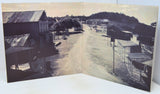 An open vintage book or photo album showing a sepia-toned street scene with buildings, trees, and a hill in the background, photographed against a plain light background.