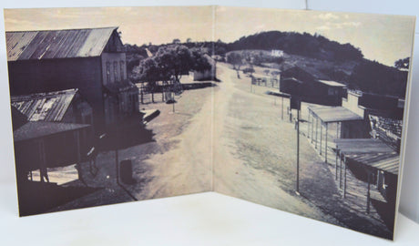 An open vintage book or photo album showing a sepia-toned street scene with buildings, trees, and a hill in the background, photographed against a plain light background.