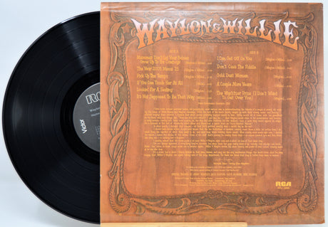 A vinyl record partially pulled from its printed sleeve, showing the back cover artwork. The black record has a visible RCA label, and the sleeve is tan and brown with ornate border graphics and the text 'Waylon & Willie' at the top.
